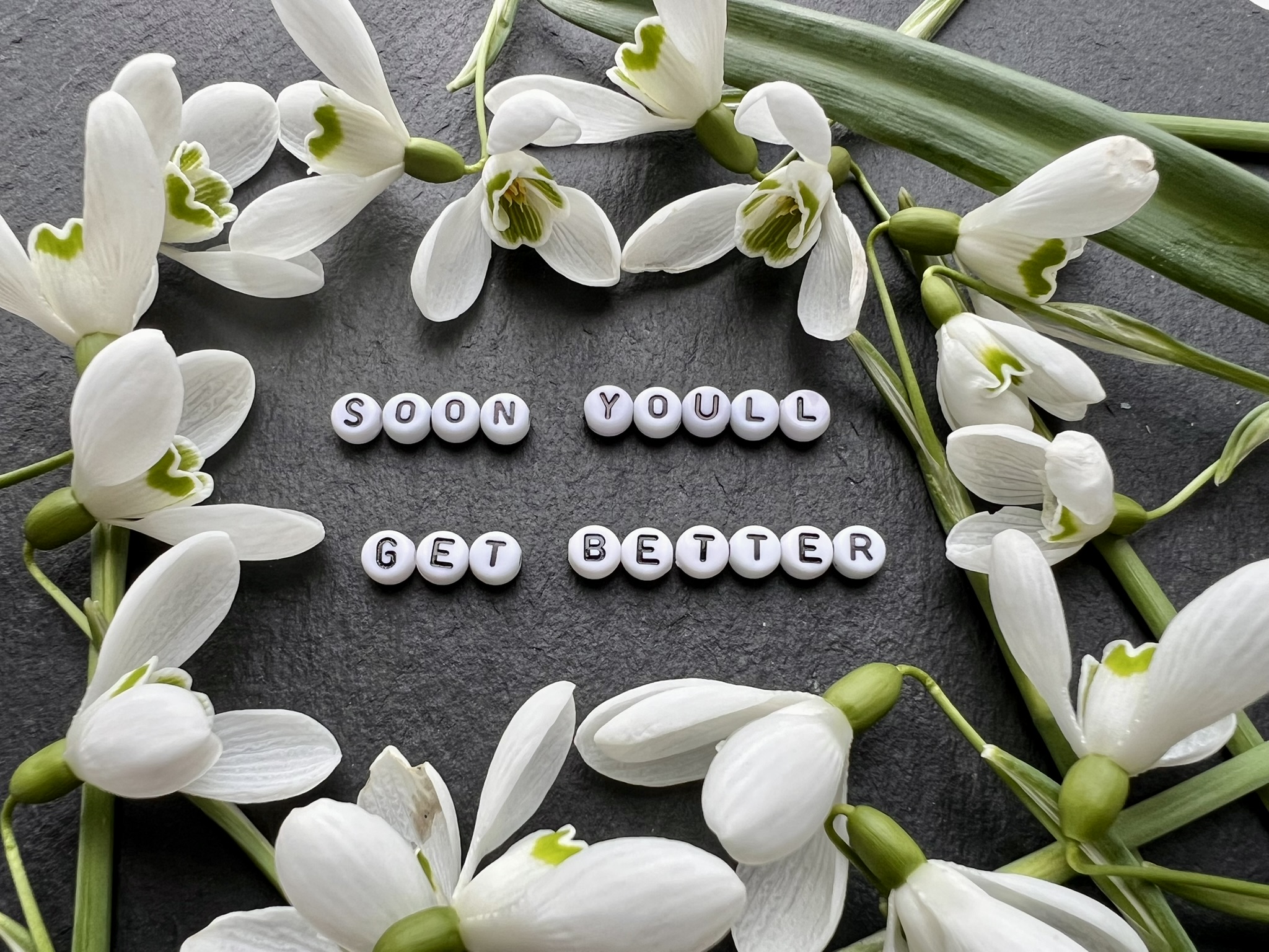 A heartfelt "get well" message on dark slate, featuring white letter beads that spell out "SOON YOU'LL GET BETTER," a direct reference to Taylor Swift's emotional song from her album Lover. The text is framed by delicate, white snowdrop flowers, symbolizing hope and resilience.
