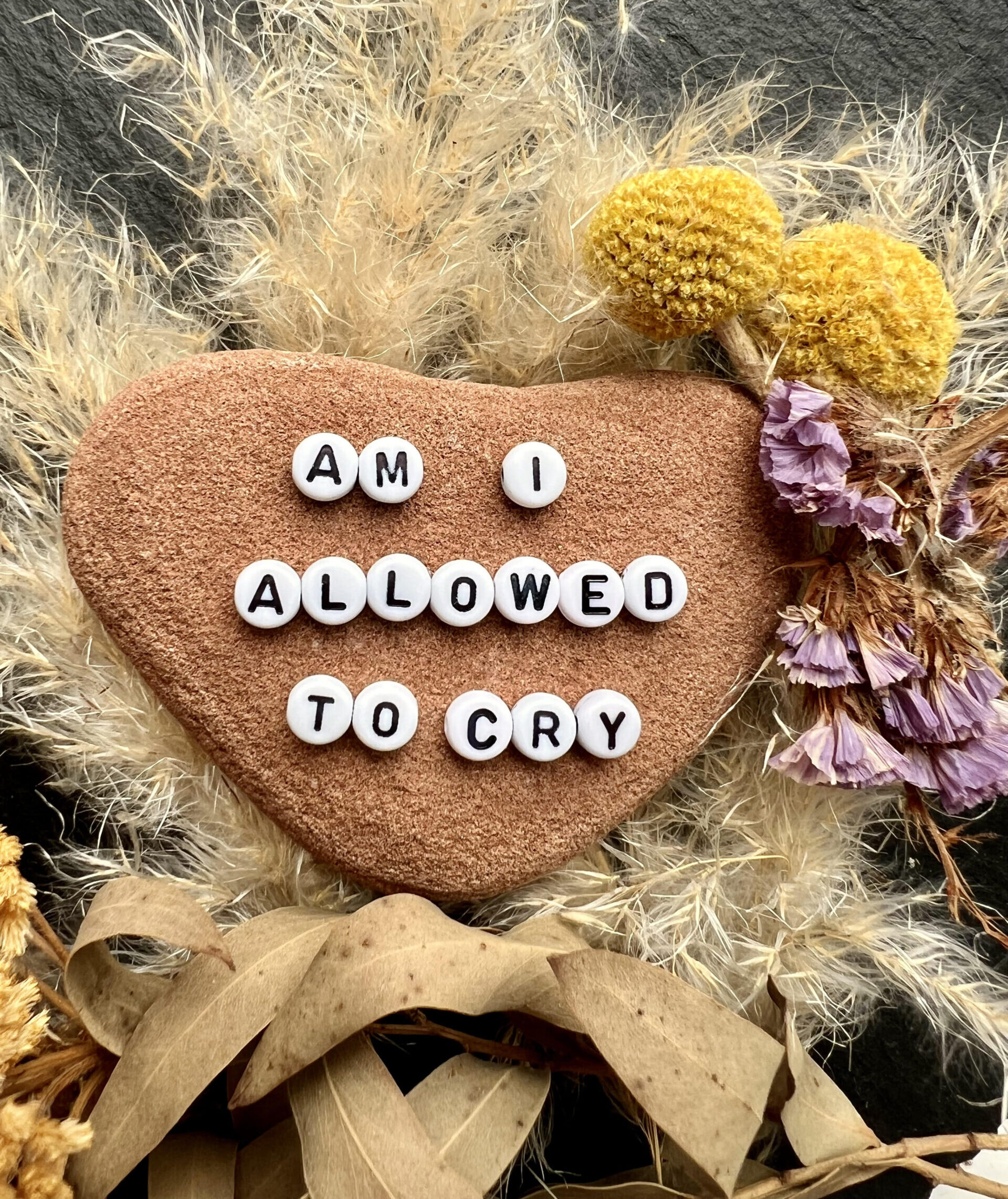 A heart-shaped stone featuring the message 'AM I ALLOWED TO CRY' in white letter beads, accented with dried flowers and grass